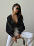 A model is sitting on a stool wearing a plunging black blouse with volume sleeves, white high-waisted pants, futuristic sunglasses and minimalist jewelry