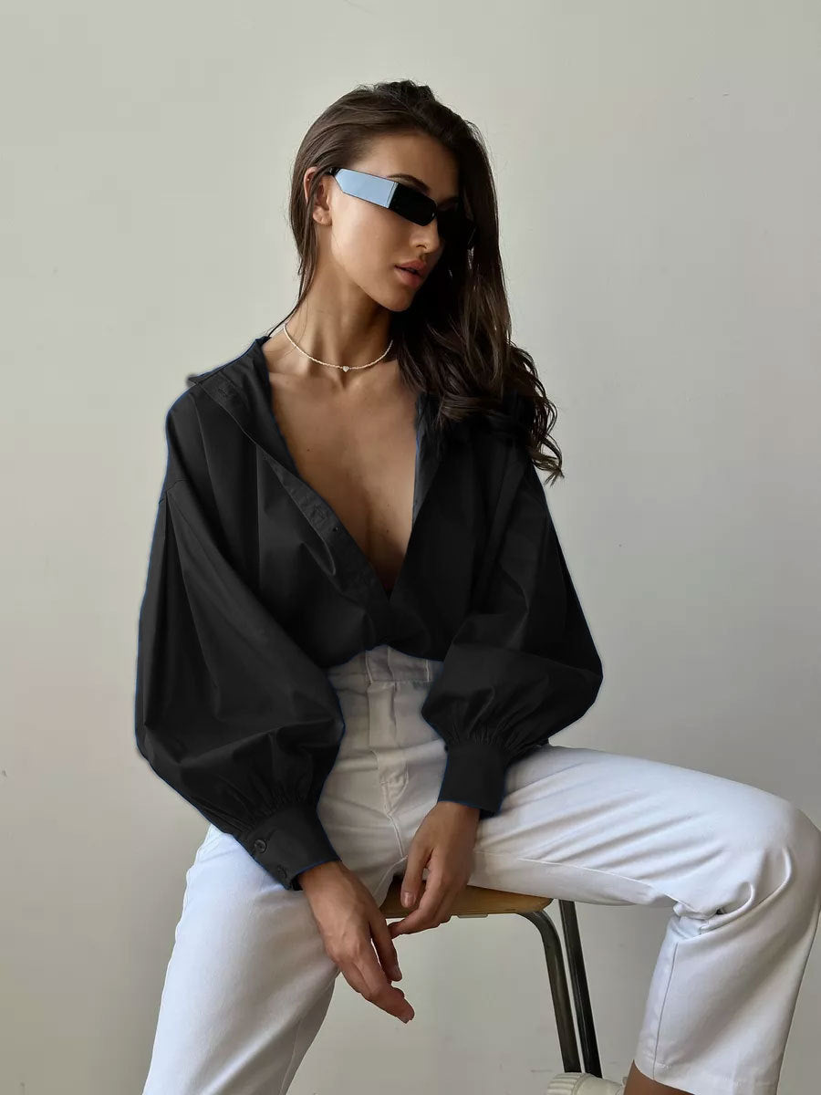 A model is sitting on a stool wearing a plunging black blouse with volume sleeves, white high-waisted pants, futuristic sunglasses and minimalist jewelry