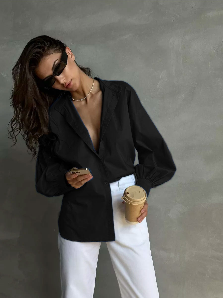 A woman is checking her phone while holding coffee, wearing a deep-v black blouse, white trousers, rectangular sunglasses and dainty gold chain necklace