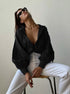 A model is posing with head tilted back on stool, wearing black oversized shirt with dramatic collar, white pants, black sunglasses and gold chain