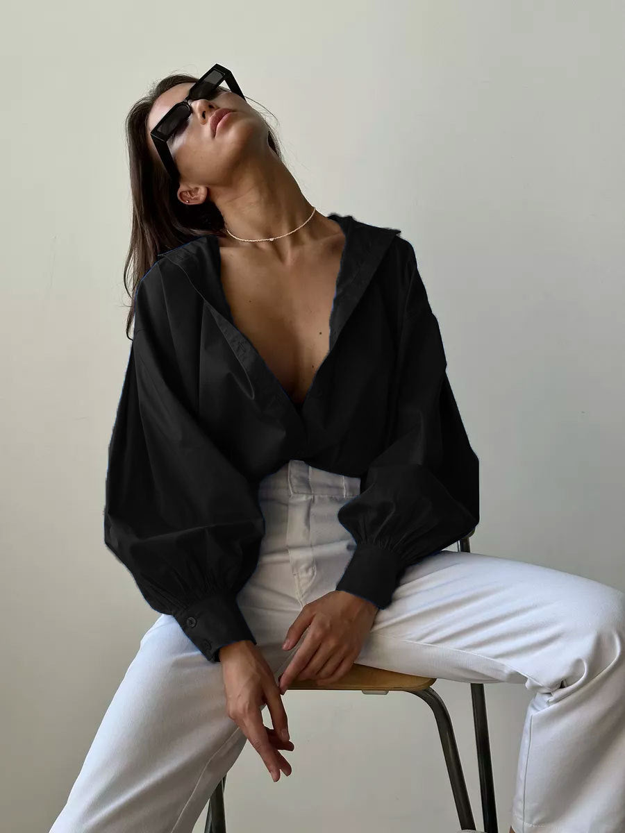 A model is posing with head tilted back on stool, wearing black oversized shirt with dramatic collar, white pants, black sunglasses and gold chain