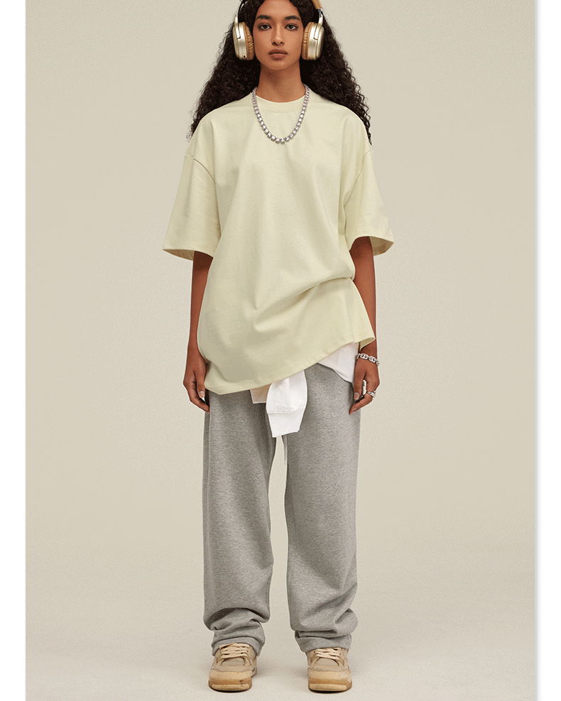 Woman wearing cream oversized t-shirt with light gray sweatpants, white headphones, silver chain necklace and baseball cap against neutral background