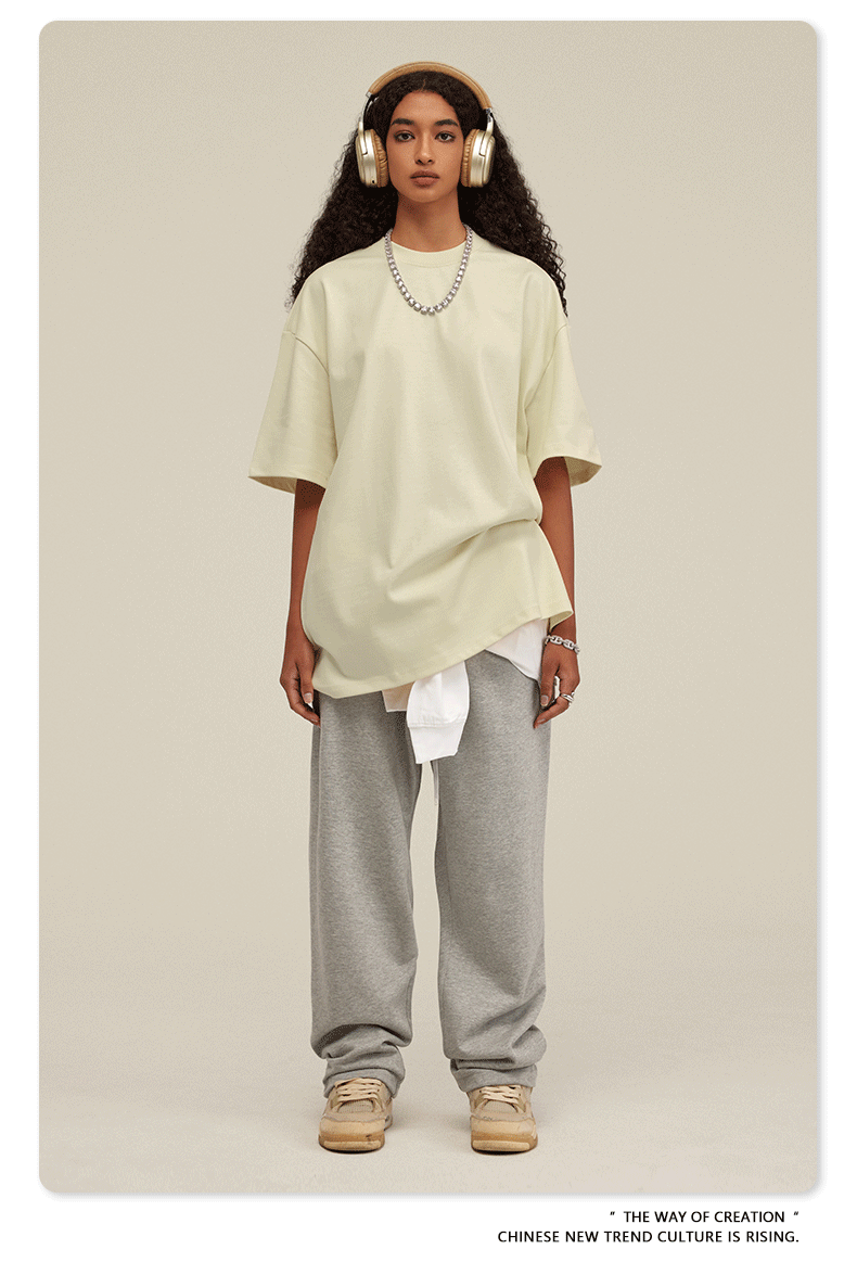Woman wearing cream oversized t-shirt with light gray sweatpants, white headphones, silver chain necklace and baseball cap against neutral background
