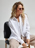 A woman wearing a white textured button-down shirt with beige pants, sitting on a black chair against white background, with wavy blonde hair and sunglasses
