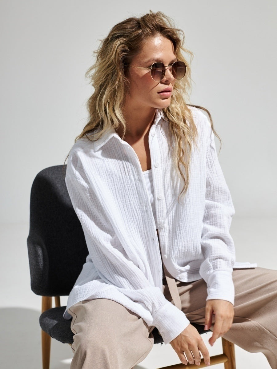 A woman wearing a white textured button-down shirt with beige pants, sitting on a black chair against white background, with wavy blonde hair and sunglasses