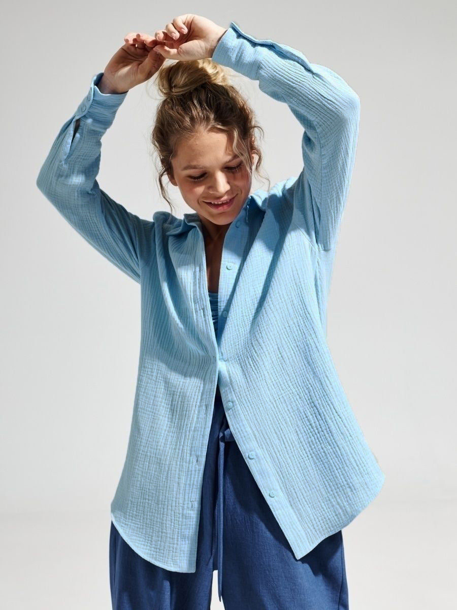 A woman wearing a light blue textured button-down shirt over navy pants, smiling while adjusting her hair in a bun pose against white backdrop