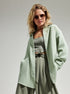 A woman wearing a mint green textured button-down shirt with sage pants and sunglasses, hands in pockets posing against white background