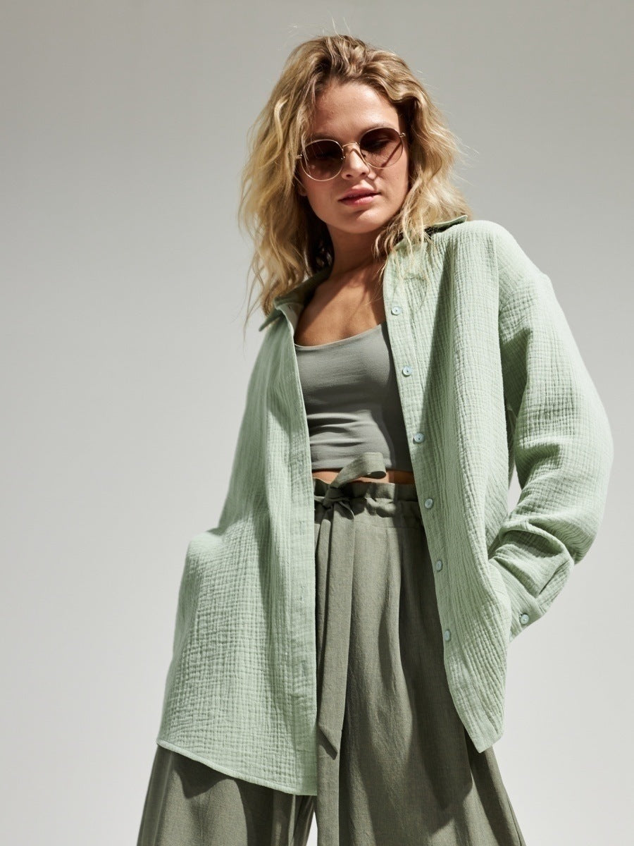 A woman wearing a mint green textured button-down shirt with sage pants and sunglasses, hands in pockets posing against white background
