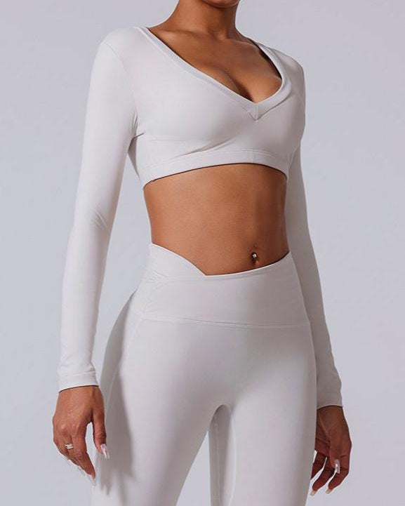 A model is wearing an all-white yoga set with V-neck long sleeve crop top and matching high-waisted leggings against neutral gray background