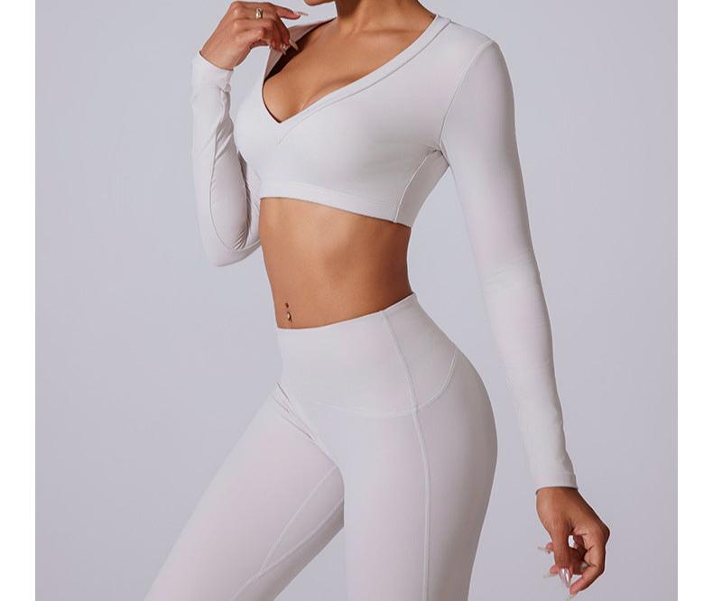 A model is posing with hand near face wearing white long sleeve V-neck crop top and matching high-waisted leggings against neutral backdrop