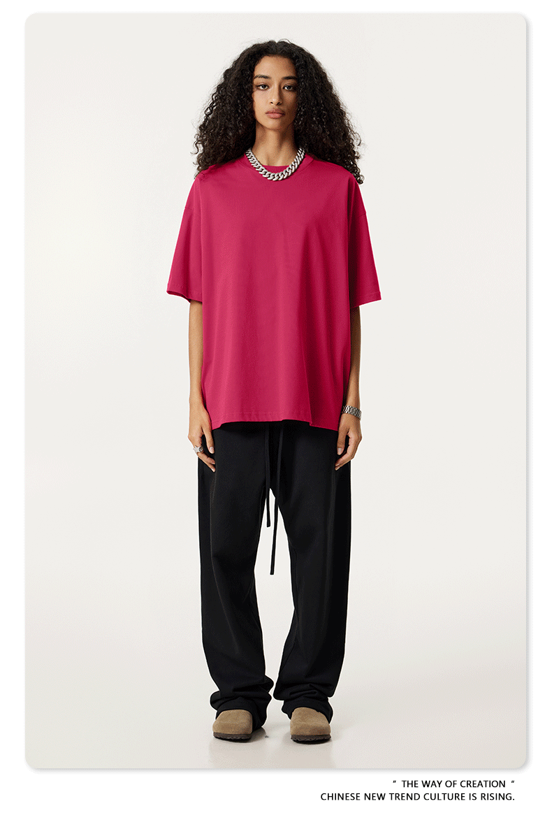Female model wearing hot pink oversized t-shirt with black relaxed pants, silver chain necklace and beige shoes against clean white background