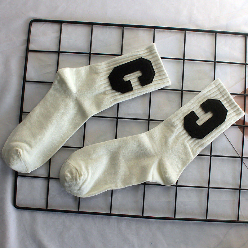 A pair of cream varsity-style crew socks with black textured "C" patches is displayed on a black wire grid against a neutral background
