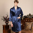 Man in royal blue satin robe with white piping. Close-up shows hand in pocket posture and classic shawl collar design against neutral background