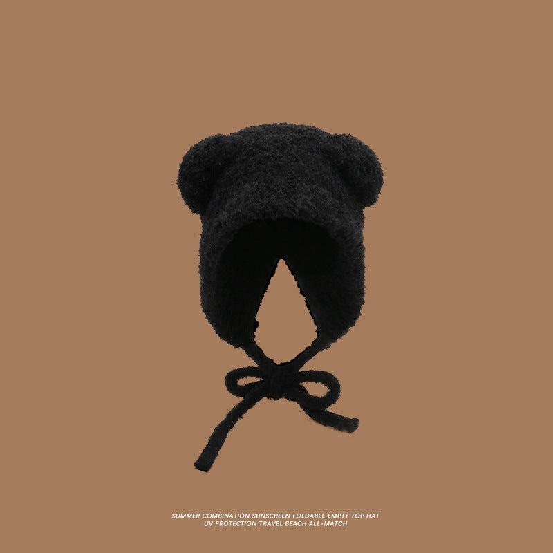 A black teddy bear bonnet with ear details and chin ties displayed against brown background, showcasing plush material construction
