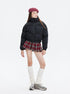 A woman wears a black puffer jacket with a red plaid pleated skirt, white knee-high socks, and gray sneakers, posing against white