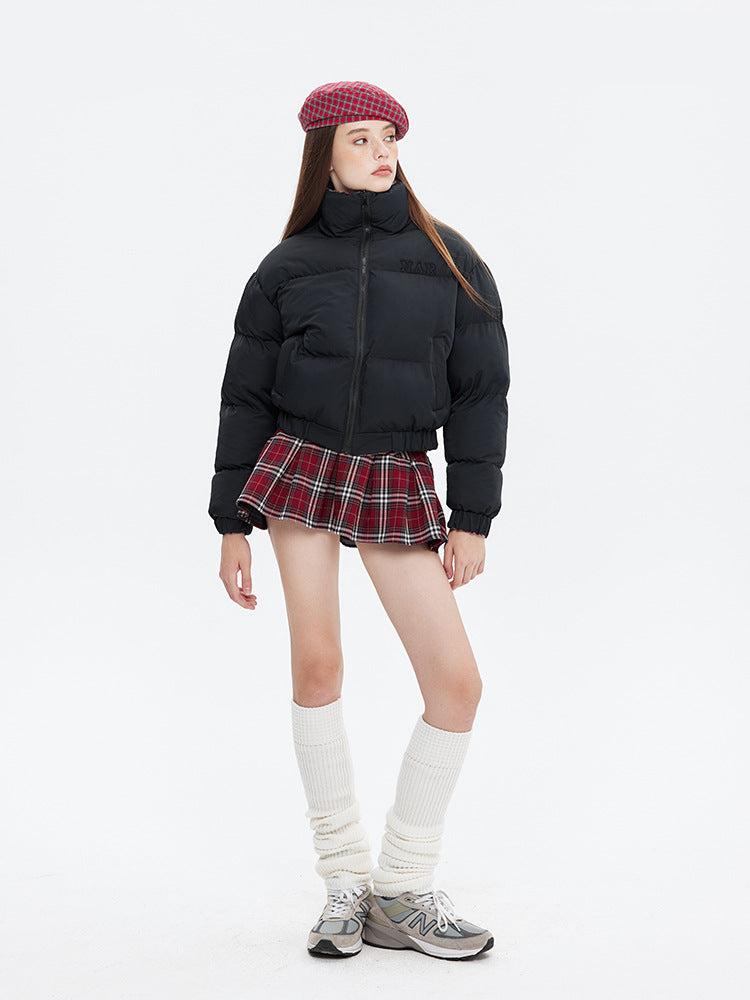 A woman wears a black puffer jacket with a red plaid pleated skirt, white knee-high socks, and gray sneakers, posing against white