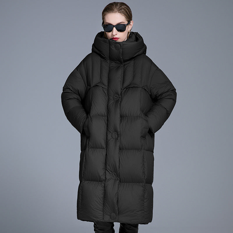 A woman wearing a black oversized quilted puffer coat with hands in pockets, black sunglasses and red lipstick, standing against gray background
