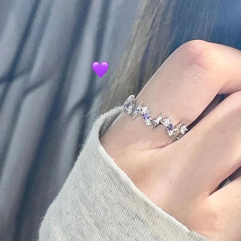 A person wearing silver ring with star and crystal design featuring purple accents, shown against blue fabric with purple heart emoji overlay