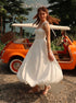 Woman in a white dress standing next to an orange vintage car with a scenic background.