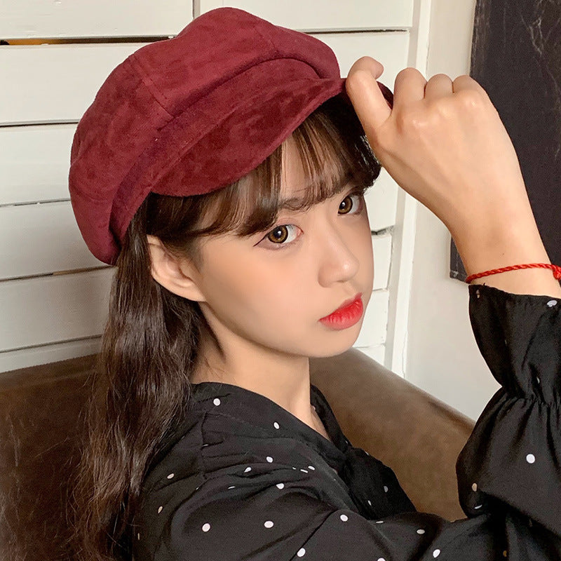 A woman wearing a burgundy suede newsboy cap, adjusting the brim while posing against white wooden wall panels