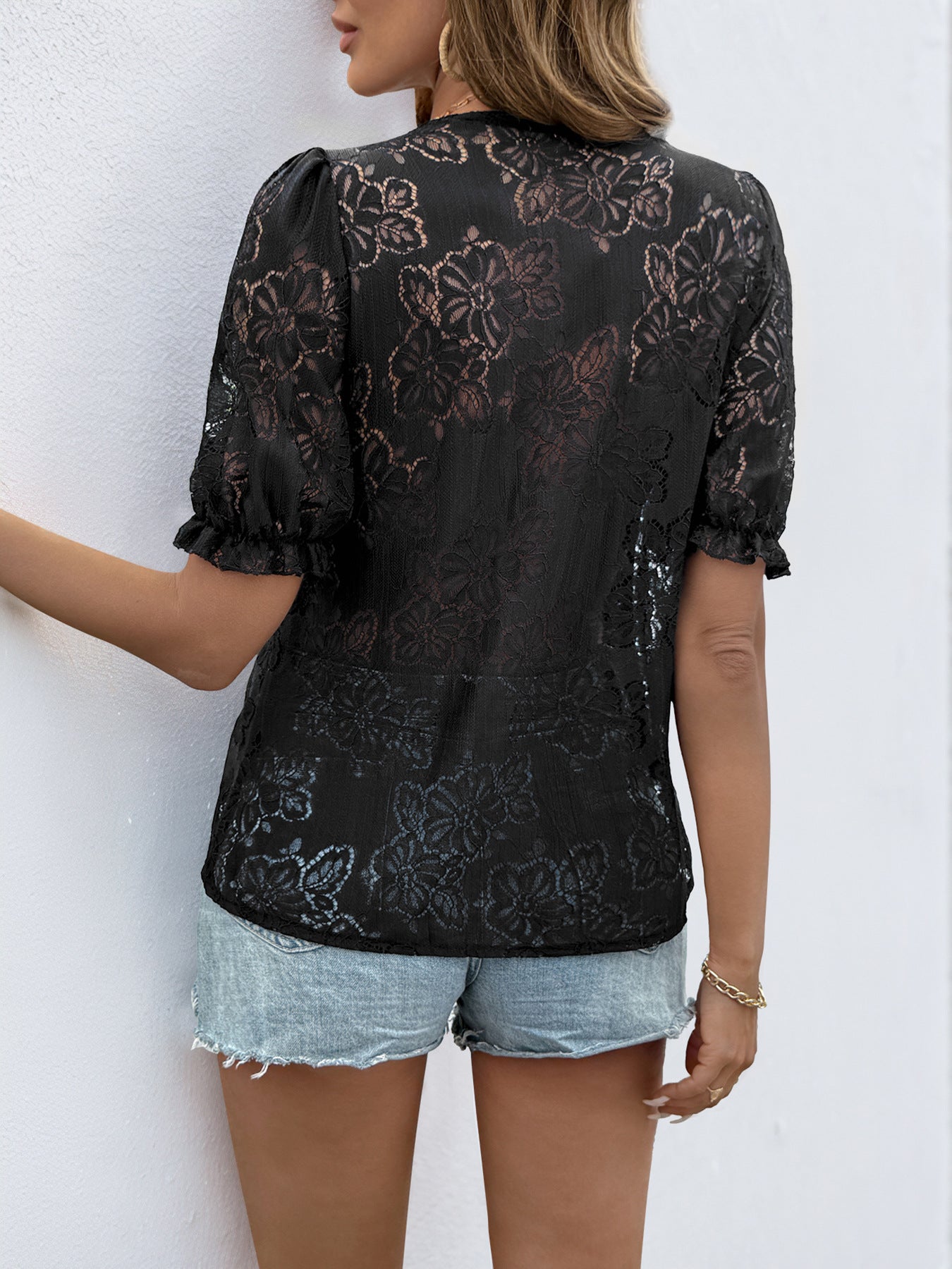 Back view of a women wearing sheer black lace blouse featuring floral embroidery and ruffled sleeves, paired with denim shorts.