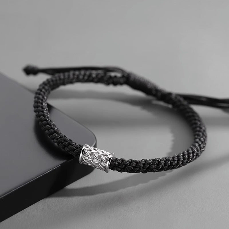 Close-up of a black braided bracelet with a silver cylindrical charm, placed on a dark surface next to a black smartphone.
