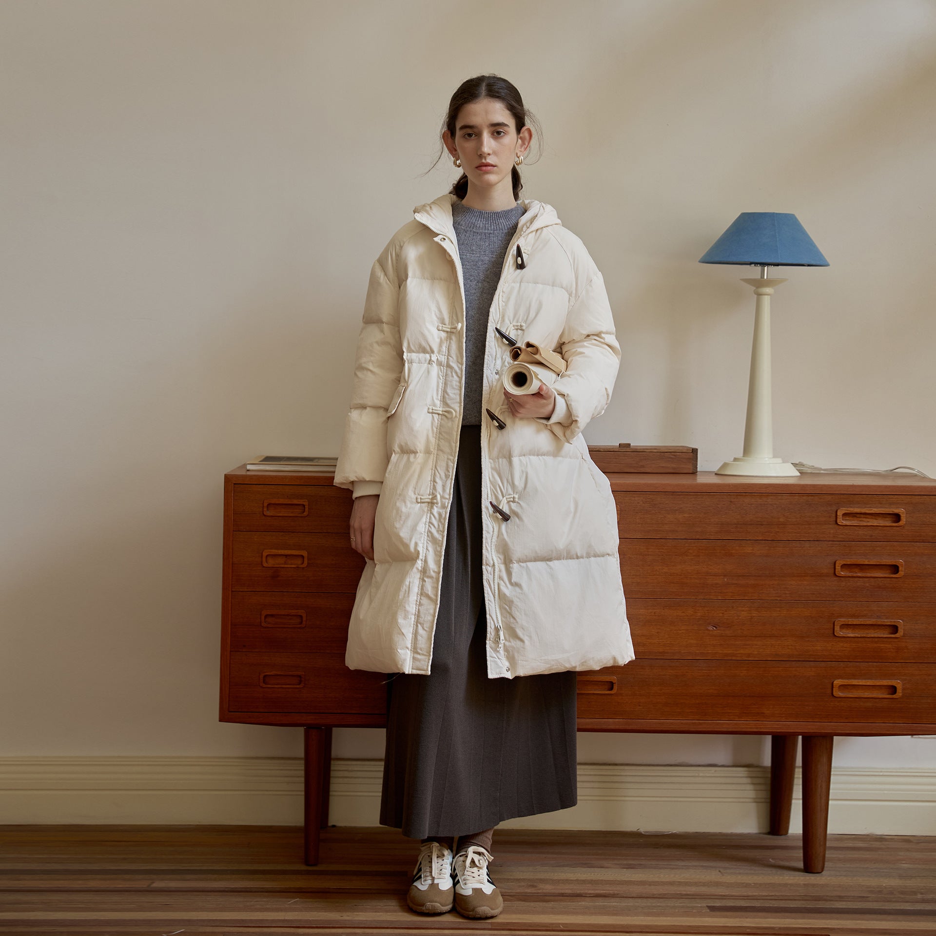 White down duffle coat styled with gray sweater dress and vintage sneakers against wooden dresser