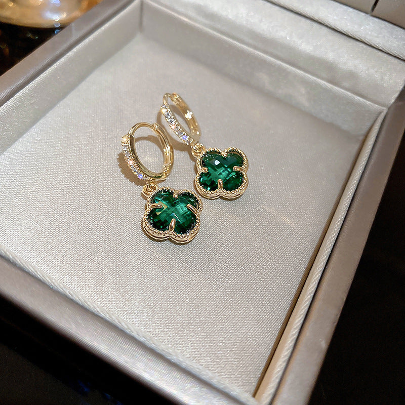Gold drop earrings with emerald green clover-shaped stones and diamond accents displayed in white jewelry box against neutral background