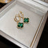 Gold drop earrings with emerald green clover-shaped stones and diamond accents displayed in white jewelry box against neutral background