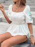 Woman sitting on steps, wearing a white smocked peplum blouse with embroidered puff sleeves, accessorized with layered necklaces.