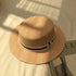 A khaki straw boater hat with a black-and-white ribbon band and silver charm, placed on a white bed under soft natural light.