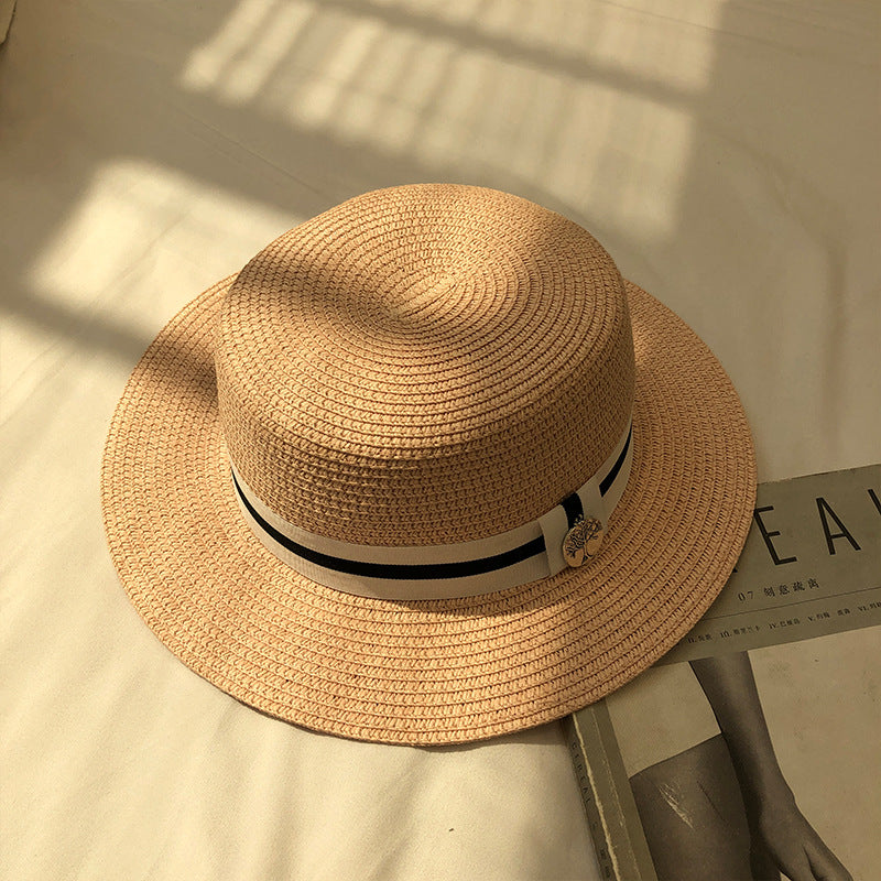 A khaki straw boater hat with a black-and-white ribbon band and silver charm, placed on a white bed under soft natural light.
