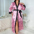 Pink satin robe with black trim, printed with handwritten hearts and "love" text, worn open with side slit on white textured bed