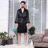 Full body front view of man wearing tied black silk robe standing barefoot on white carpet with decorative plants and tweed furniture in background