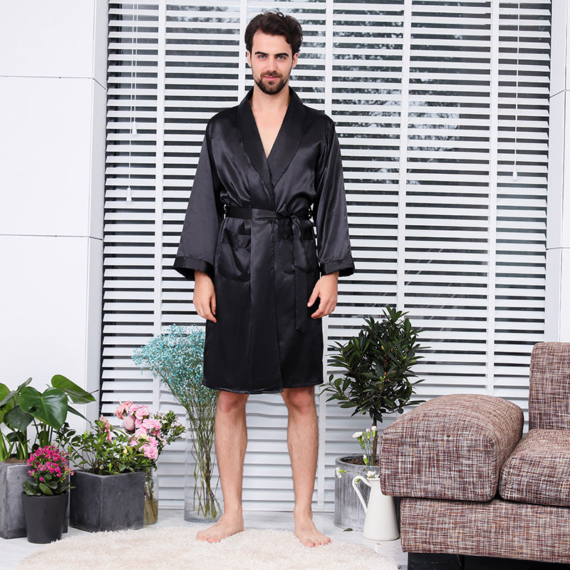 Full body front view of man wearing tied black silk robe standing barefoot on white carpet with decorative plants and tweed furniture in background