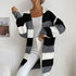 A woman in black, white and taupe striped open cardigan paired with white tank top and white jeans standing against white wall