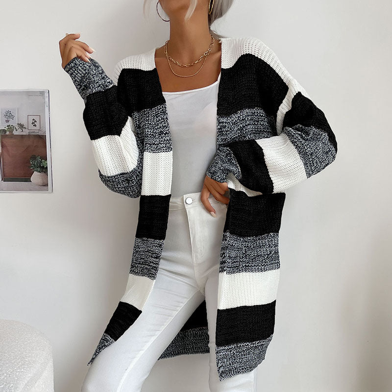 A woman in black, white and taupe striped open cardigan paired with white tank top and white jeans standing against white wall
