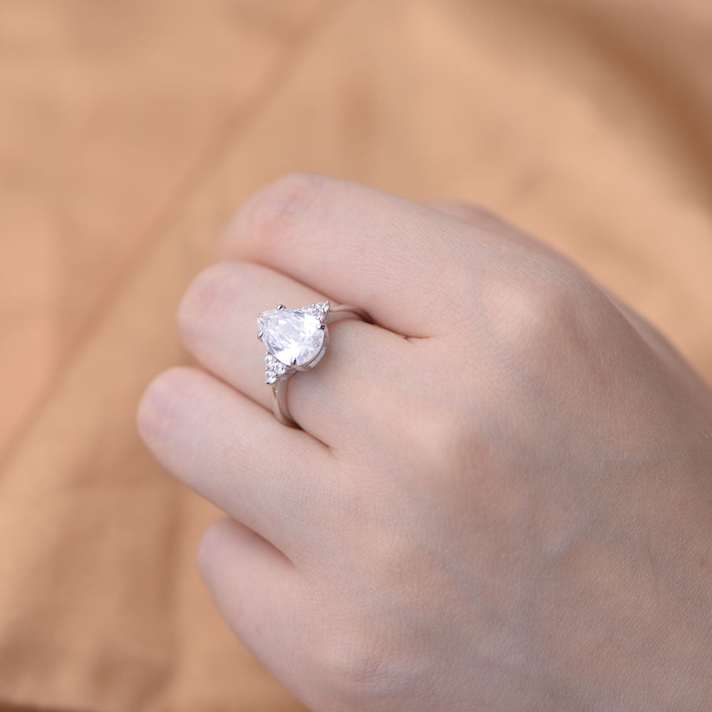 A woman wearing a silver pear-cut diamond engagement ring on her finger, posed against a warm beige background showing elegant side profile