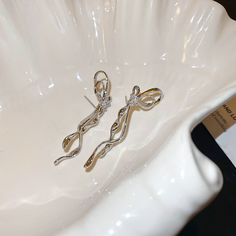 Minimalist silver bow earrings with curved ribbons shown on white ceramic display, highlighting clean lines