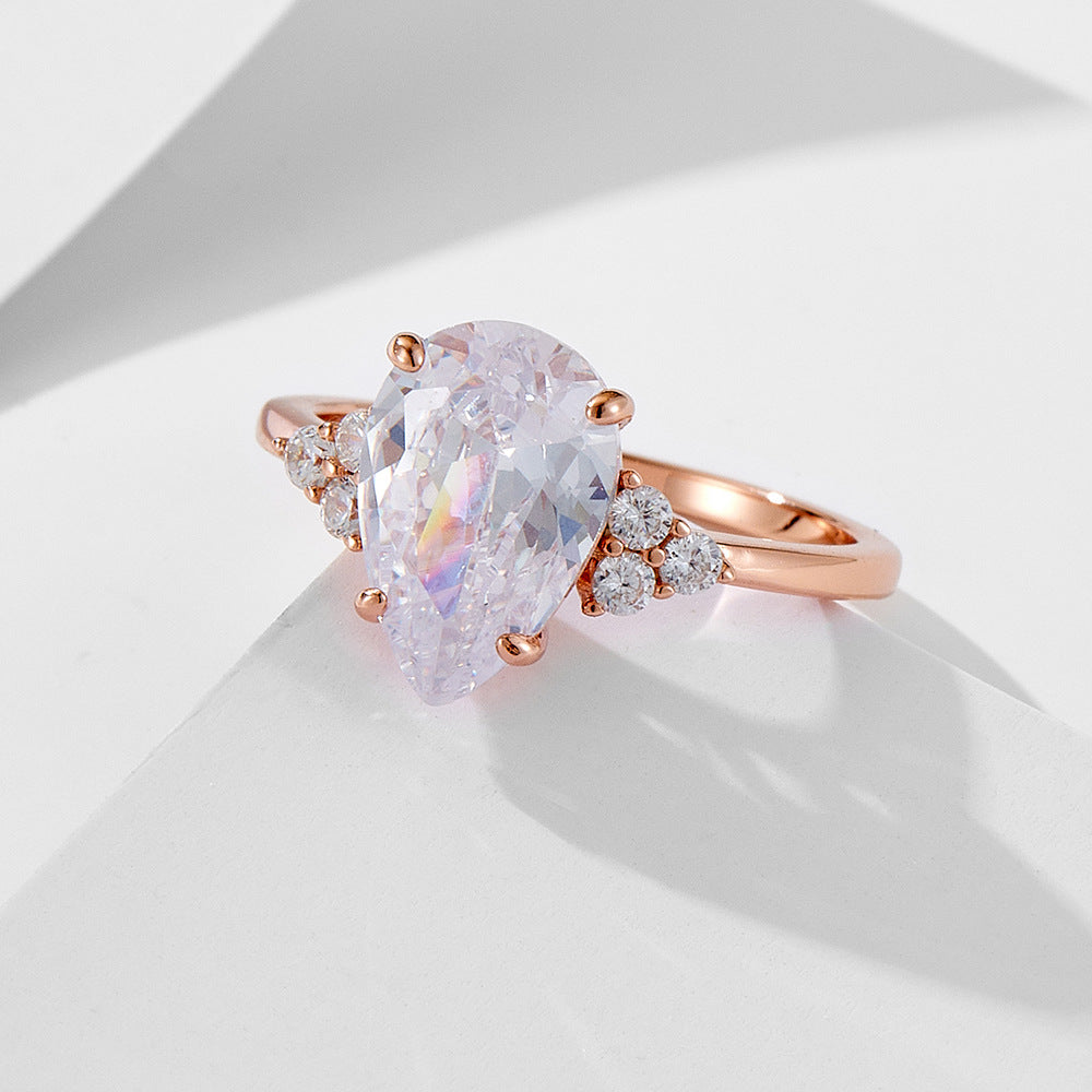 A rose gold engagement ring with large pear-cut crystal and cluster diamond accents on both sides, photographed on white geometric surface