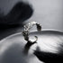 A unique silver ring with an abstract organic texture displayed on a reflective plate with black feathers in the background.