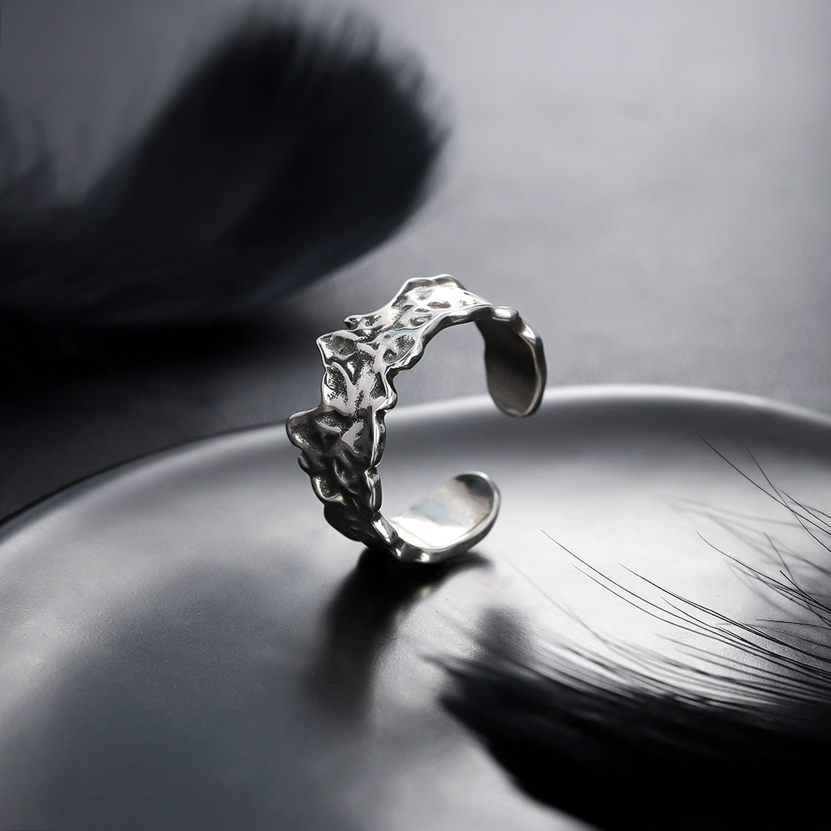 A unique silver ring with an abstract organic texture displayed on a reflective plate with black feathers in the background.