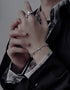 A person wearing a silver chain bracelet with a cross pendant, paired with a silver statement ring, dressed in a dark outfit with a patterned shirt.