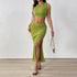 A stylish woman walking in a green ribbed two-piece outfit, accessorized with a gold chain handbag and clear high heels, emphasizing the thigh-high slit.
