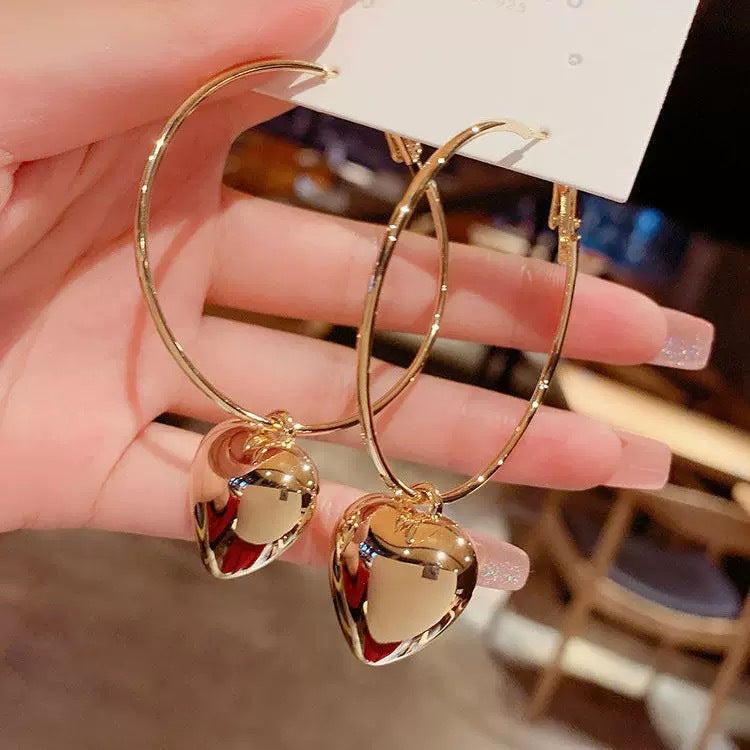 A woman holding gold hoop earrings with heart-shaped pendants attached to jewelry display card with manicured nails