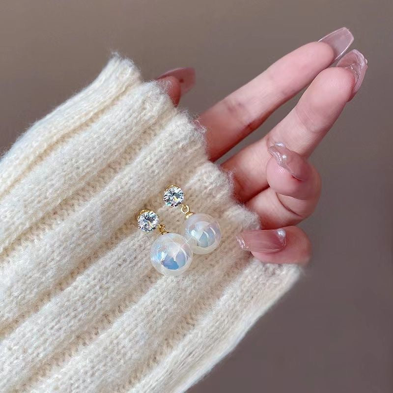 A woman's hand wearing cream sweater sleeve displaying elegant pearl drop earrings with crystal stud tops against neutral background