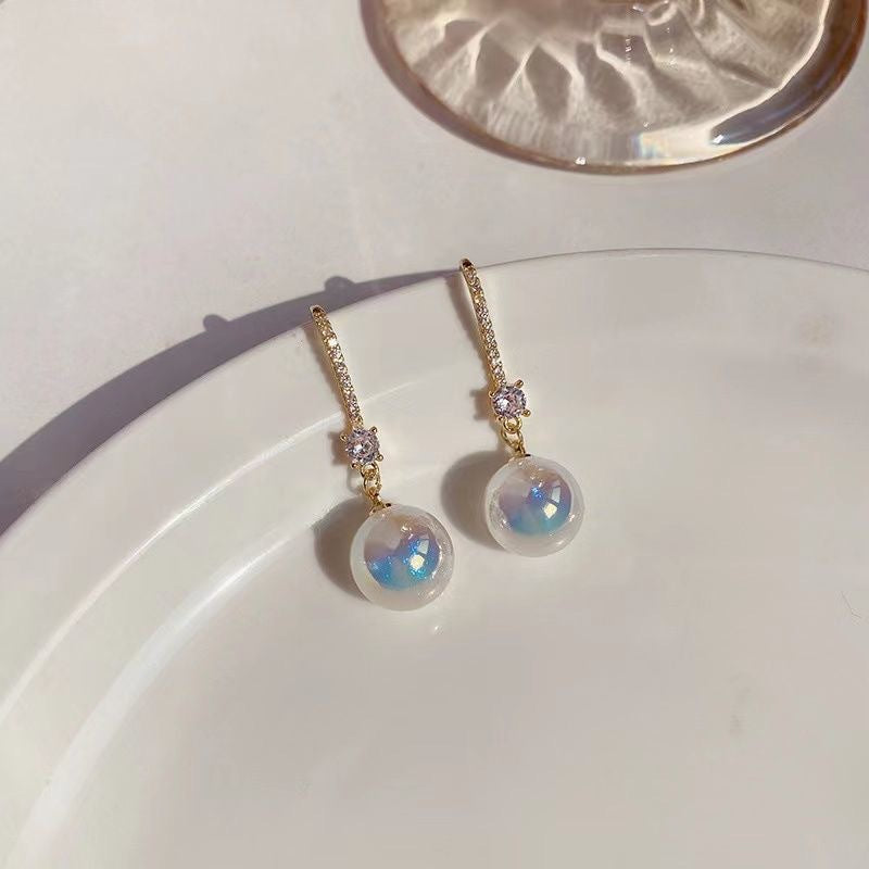 Gold earrings with crystal accents and iridescent pearl drops displayed on white ceramic plate with natural light casting shadows