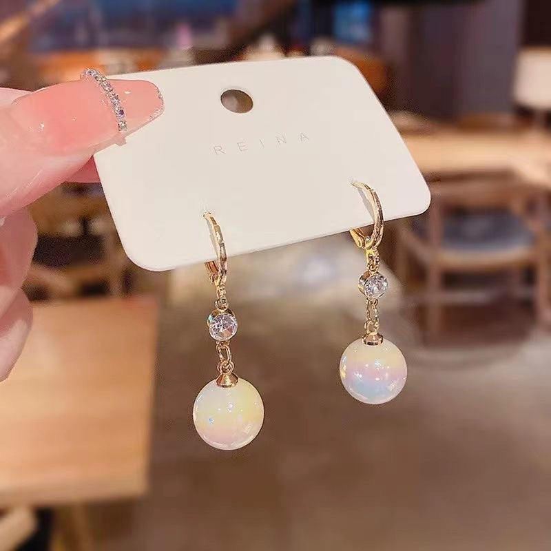 A hand holding white jewelry display card with REINA brand logo showing gold hoop earrings with crystal and iridescent pearl drops
