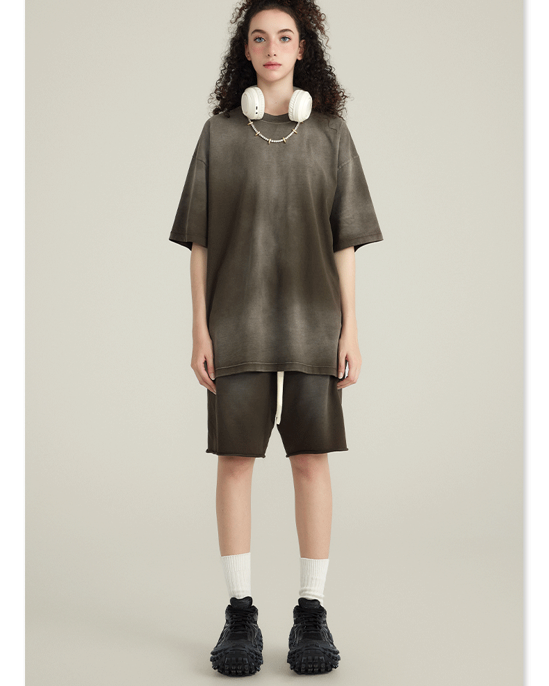 Model with curly hair wearing olive green t-shirt and matching shorts, with white headphones around neck, against cream background