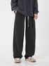 A model is wearing oversized black wide-leg sweatpants with diagonal seams and drawstring waist, paired with a dark sweatshirt and gray sneakers